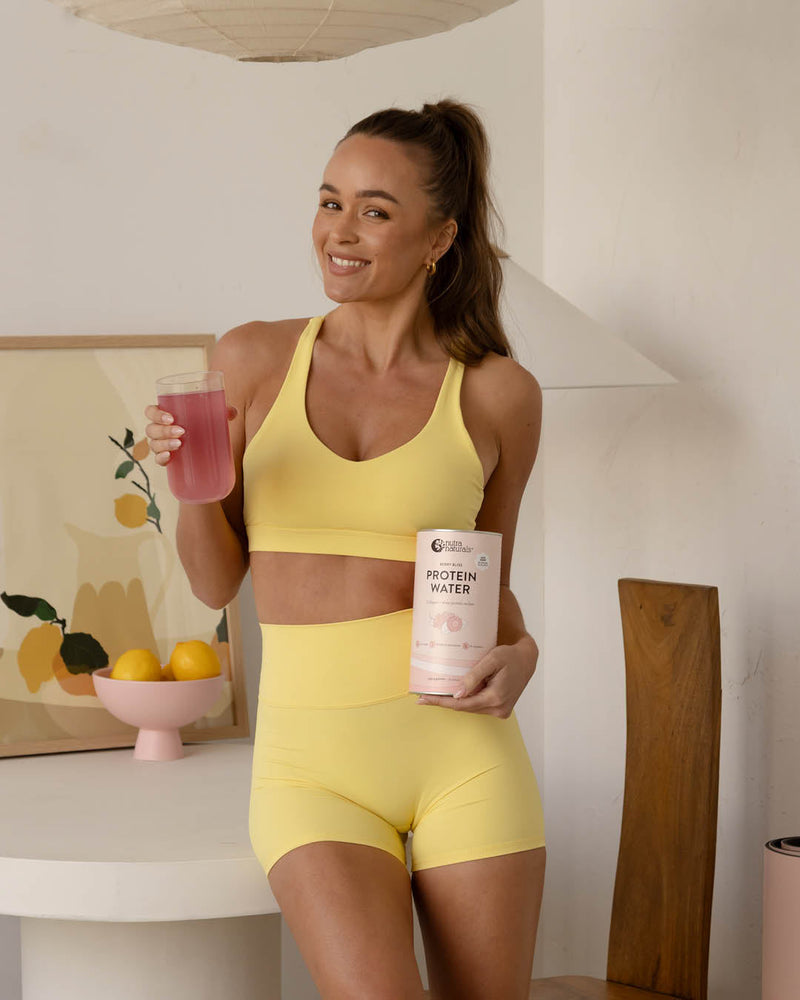 Woman in yellow athletic wear holding a protein powder container and a glass of protein water drink.