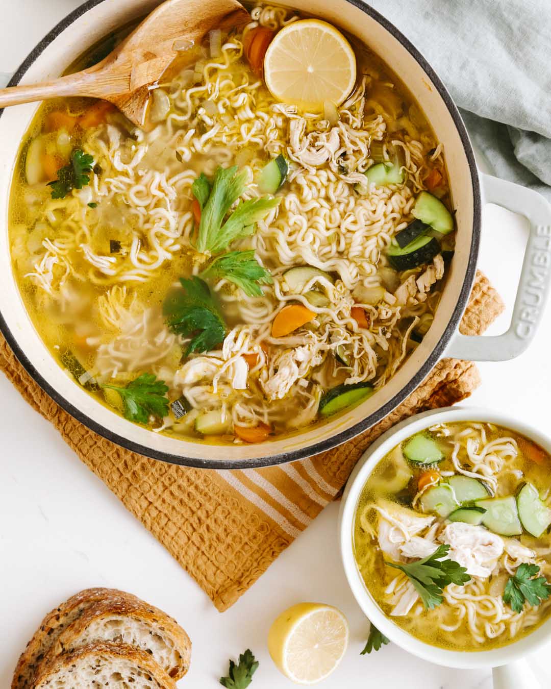 Noodle soup with vegetables in a pot, accompanied by a bowl of soup and bread slices on a white surface.