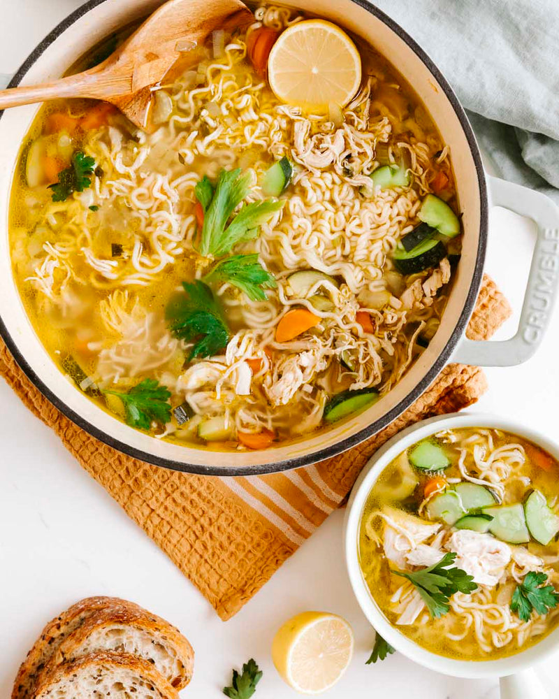 Noodle soup with vegetables in a pot, accompanied by a bowl of soup and bread slices on a white surface.