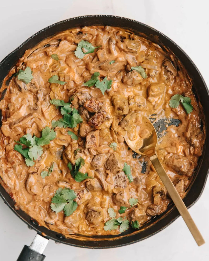 A creamy mushroom and meat curry in a black skillet, garnished with fresh cilantro and served with a gold spoon on a white background.